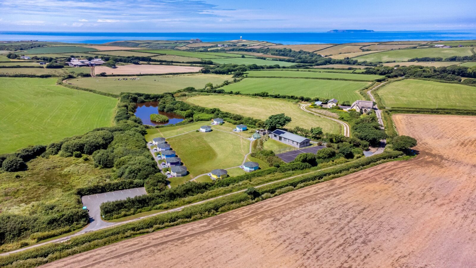 Aerial view of a countryside landscape with fields, a small lake, and a cluster of rustic cabins. The coastal sea shimmers in the background, adding a serene touch to the picturesque scene.