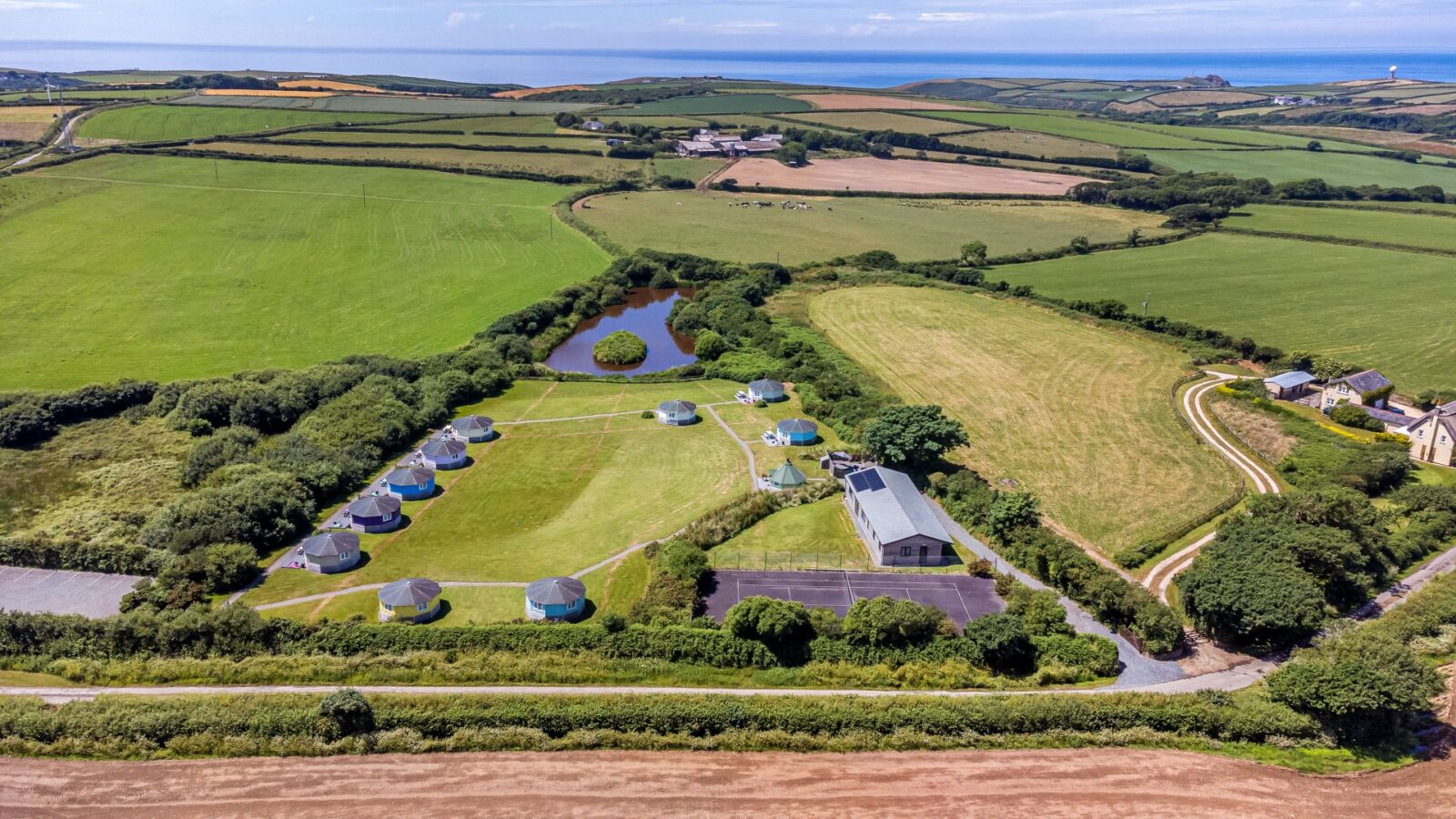 Aerial view of a coastal campsite featuring multiple tents, cozy cabins, a central building, and expansive fields nestled by the shore.