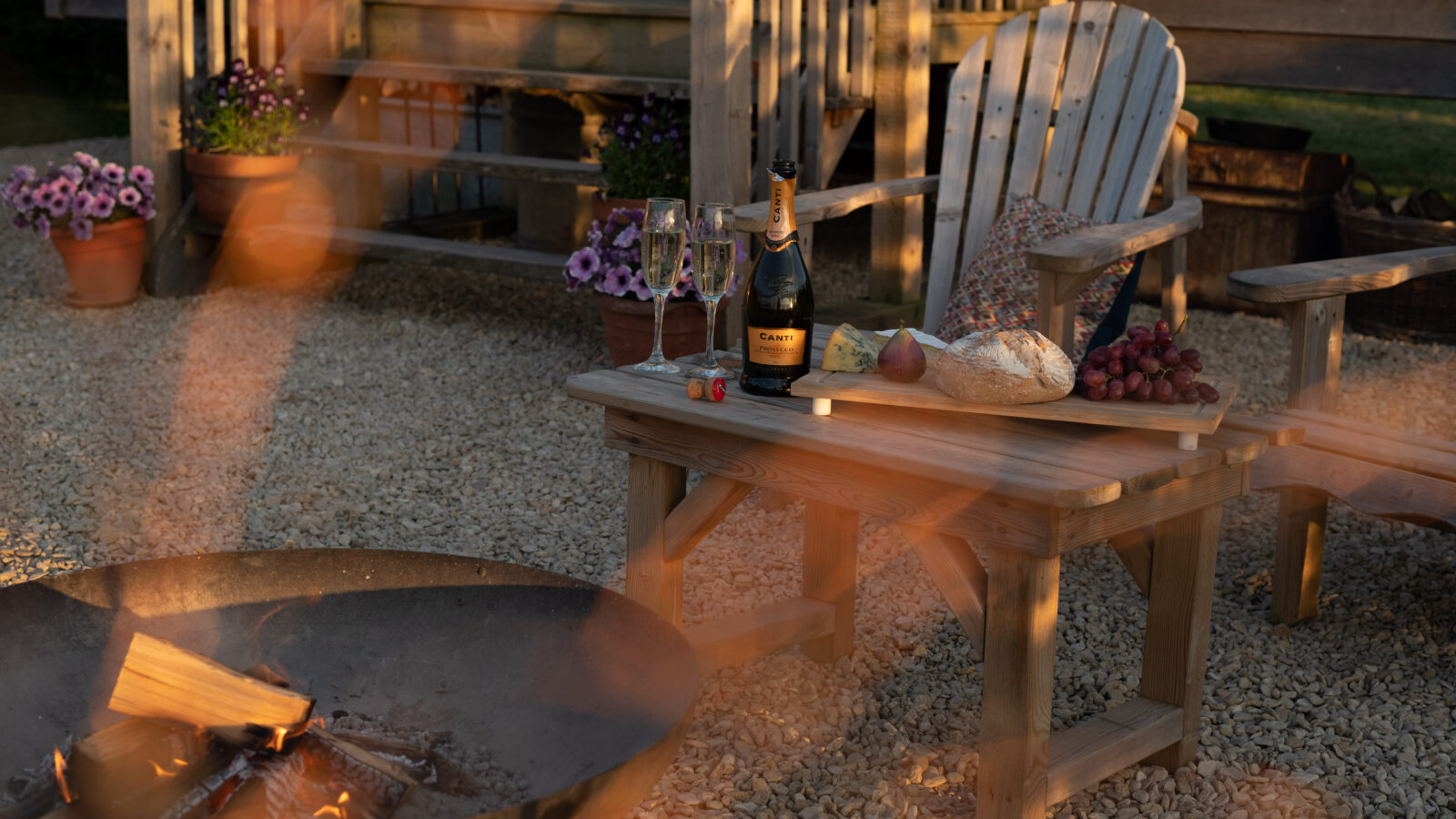 A cozy outdoor setting with a wooden table holding a bottle of sparkling wine, two filled glasses, a loaf of bread, cheese, and a bunch of grapes. Two wooden chairs sit nearby on the gravel surface. A fire pit with burning logs is in the foreground. Flowers decorate the background near charming shepherd huts.