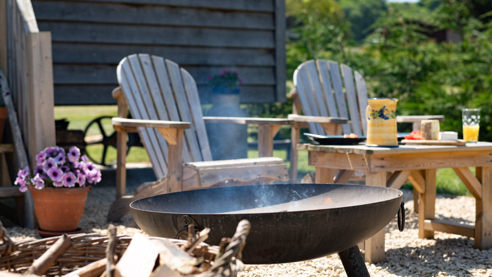 An outdoor seating area with two wooden Adirondack chairs around a fire pit filled with burning wood. A wooden table holds a vase, drinks, and food items. A woven basket with more firewood sits nearby, along with a potted plant featuring purple flowers. Nearby, a Shepherd Hut completes the cozy setup.