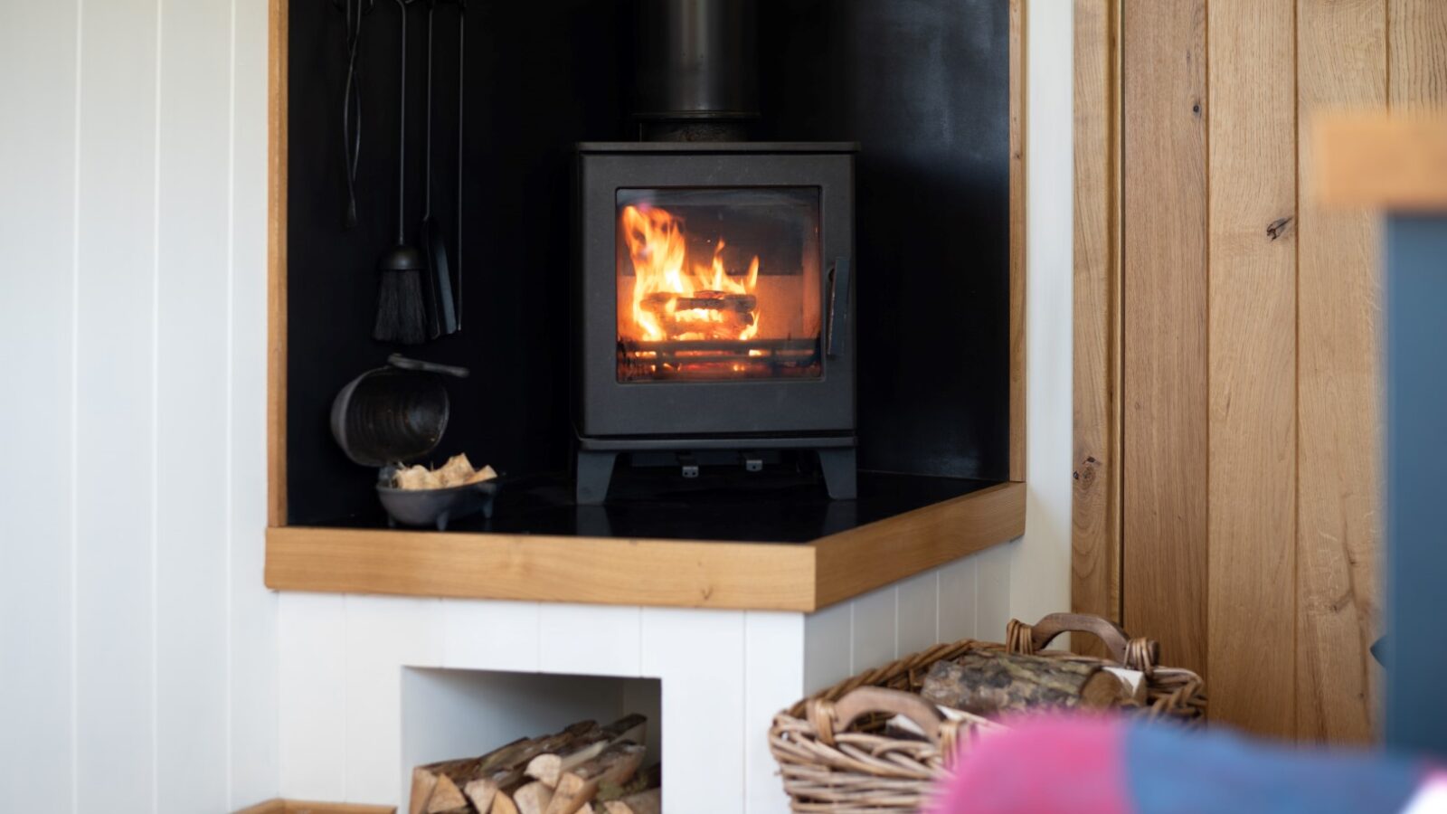 A cozy corner with a black wood-burning stove lit with a bright flame, reminiscent of traditional Shepherd Huts. The stove sits on a wooden shelf with logs stored underneath. A basket of firewood and fire tools are placed nearby. The walls are paneled in white and wood, creating a warm, inviting atmosphere.