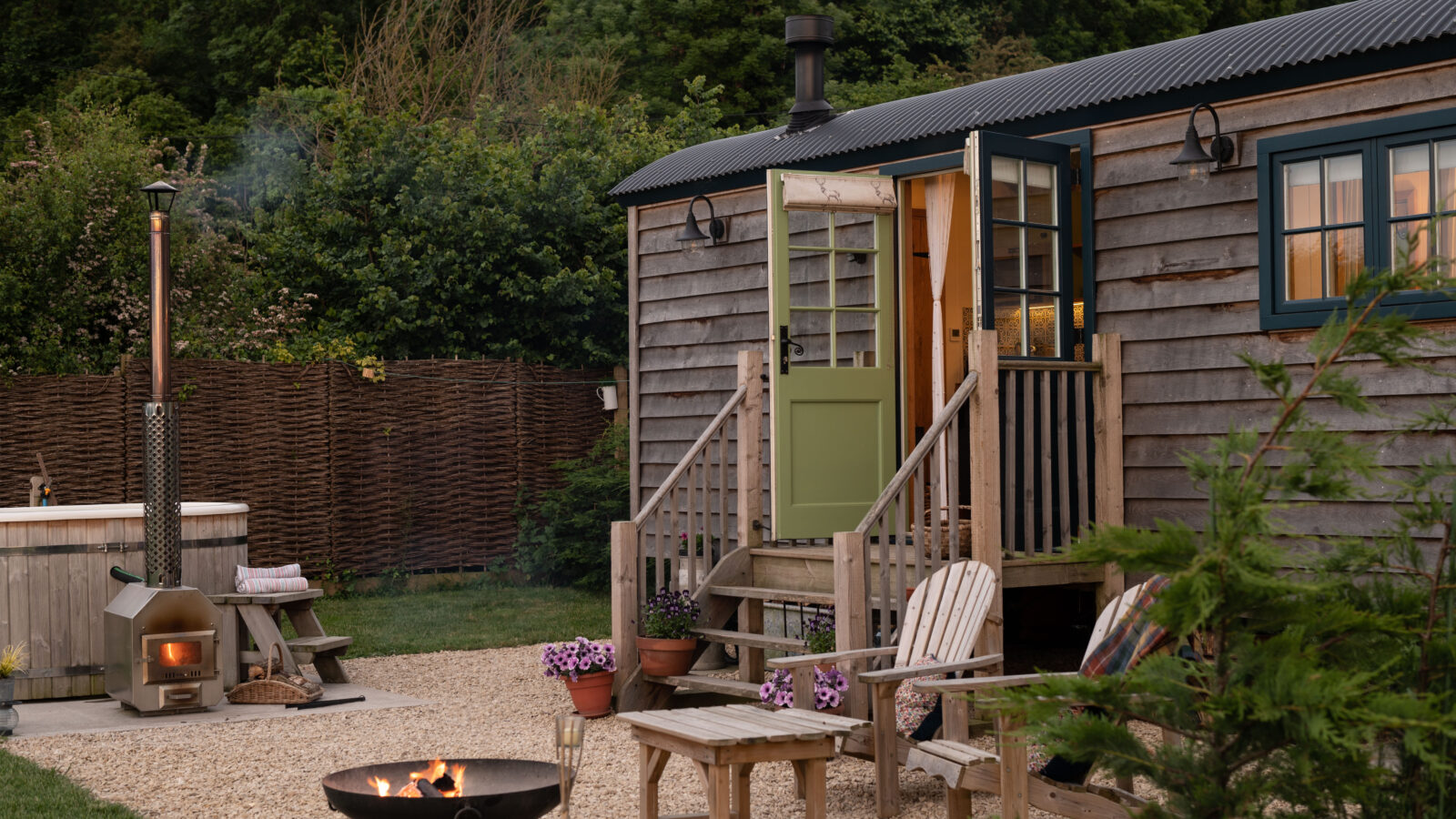 A cozy wooden cabin with a green door and steps leading to an outdoor area. The area features a fire pit with chairs arranged around it, a wood-fired oven, and a hot tub. The tranquil setting is reminiscent of shepherd huts nestled in a lush, green forest background, making it inviting and serene.