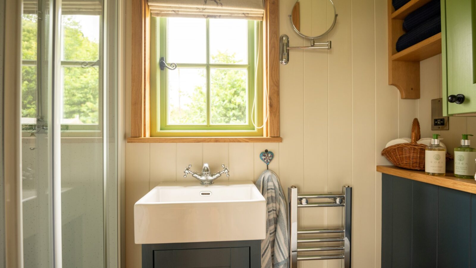 A cozy bathroom reminiscent of Shepherd Huts, with a small window above a white sink. The sink has a silver faucet and a striped towel hanging next to it. There's a shower on the left, a round mirror on the wall, a towel rack, and shelves with blue towels and toiletries on the right.