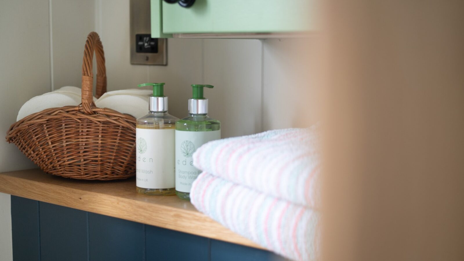 A bathroom shelf holds a wicker basket filled with white towels, two pump bottles of soap labeled 