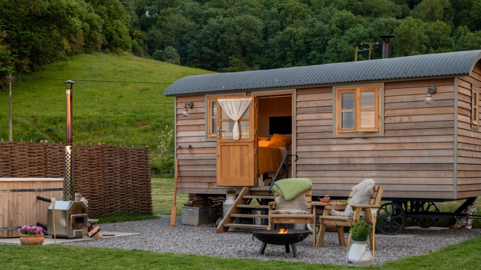 A cozy wooden tiny house on wheels with a small porch, situated on a grassy area with a lush hill behind it. The scene includes a fire pit, a chair with a green blanket, and a hot tub on the side. A friendly Collie sits contentedly by the open door, revealing the warmly lit interior reminiscent of charming shepherd huts.