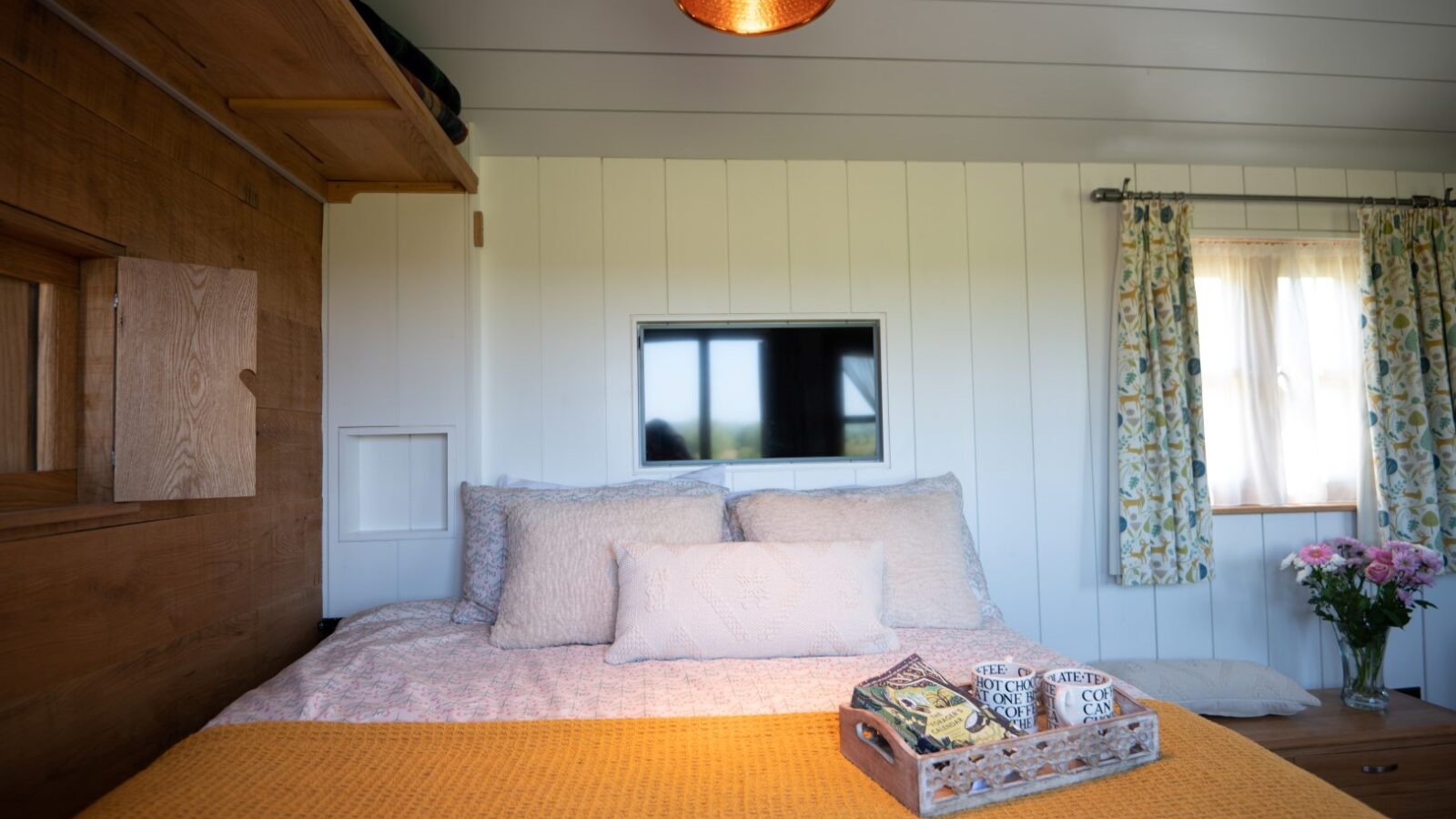 A cozy bedroom reminiscent of quaint huts, featuring a large bed with yellow and white bedding. A small window above the bed frames the scene, while a wooden lamp hangs from the ceiling. A tray with mugs rests on the bed, and pink flowers in a vase sit near a window adorned with floral curtains.