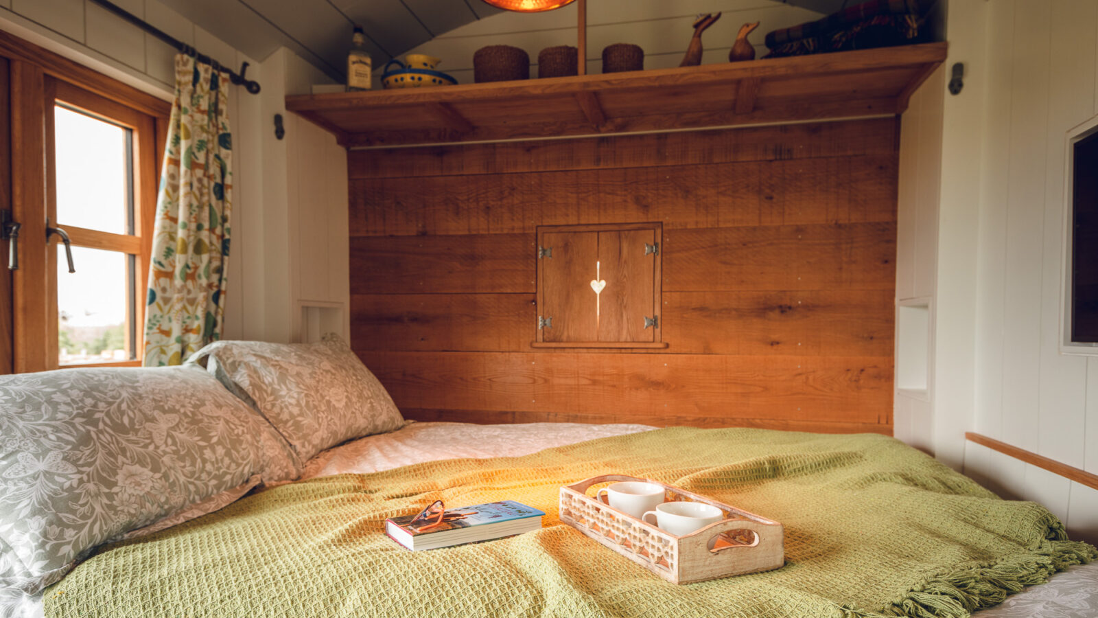 A cozy bedroom in a Collie Shepherd Hut features a double bed covered in a green knit blanket and patterned pillows. A tray with two white mugs and a book rests on the bed. Above, there's a wooden shelf with decorative items, and light filters through a window with floral curtains.