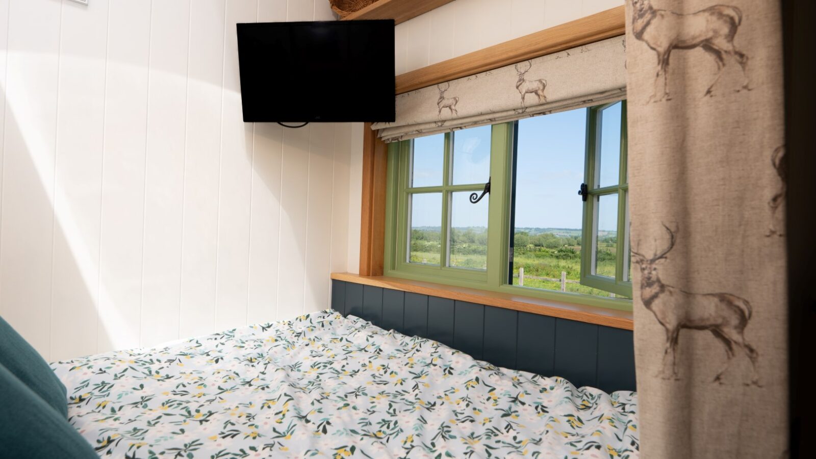 A cozy bedroom with a floral-patterned bedspread, situated near a window revealing a scenic countryside view. The white walls are adorned with wooden accents reminiscent of rustic huts. A small, mounted TV hangs above the bed, and a curtain with deer motifs partially covers the window.