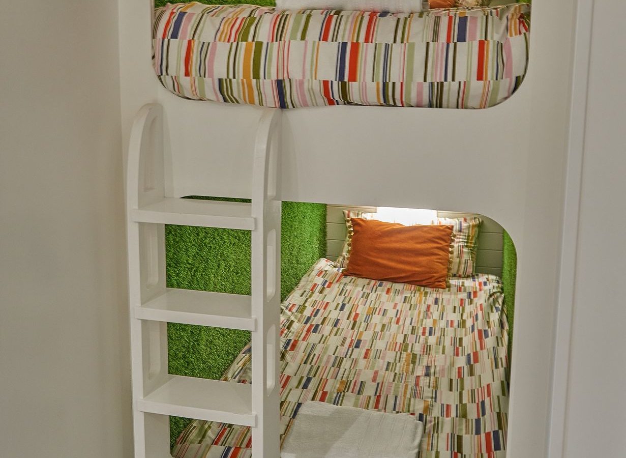 A cozy, modern bunk bed setup with colorful striped bedding and a touch of luxury reminiscent of Camlad Valley. The beds are built into a white wall, accented by a green textured background and a small rectangular window. A white ladder provides access to the top bunk.