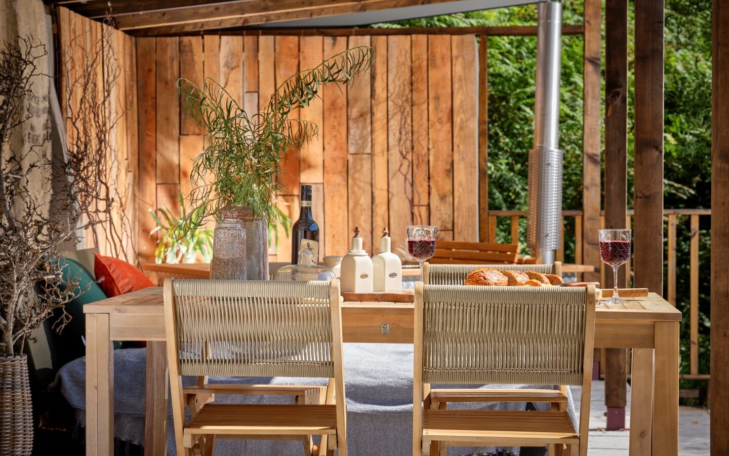 A cozy outdoor dining area in the heart of Devon features a wooden table set for two, complete with wine glasses, a bottle, and bread. Wooden chairs and vibrant greenery create a warm, inviting atmosphere under the rustic pergola at Sleepy Owl.