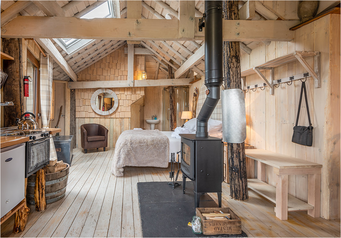 Rustic wood cabin interior with a cozy bed, wood stove, kitchenette, and armchair in Craighead Howf. Natural light spills in through skylights, enhancing the charm of this serene haven.