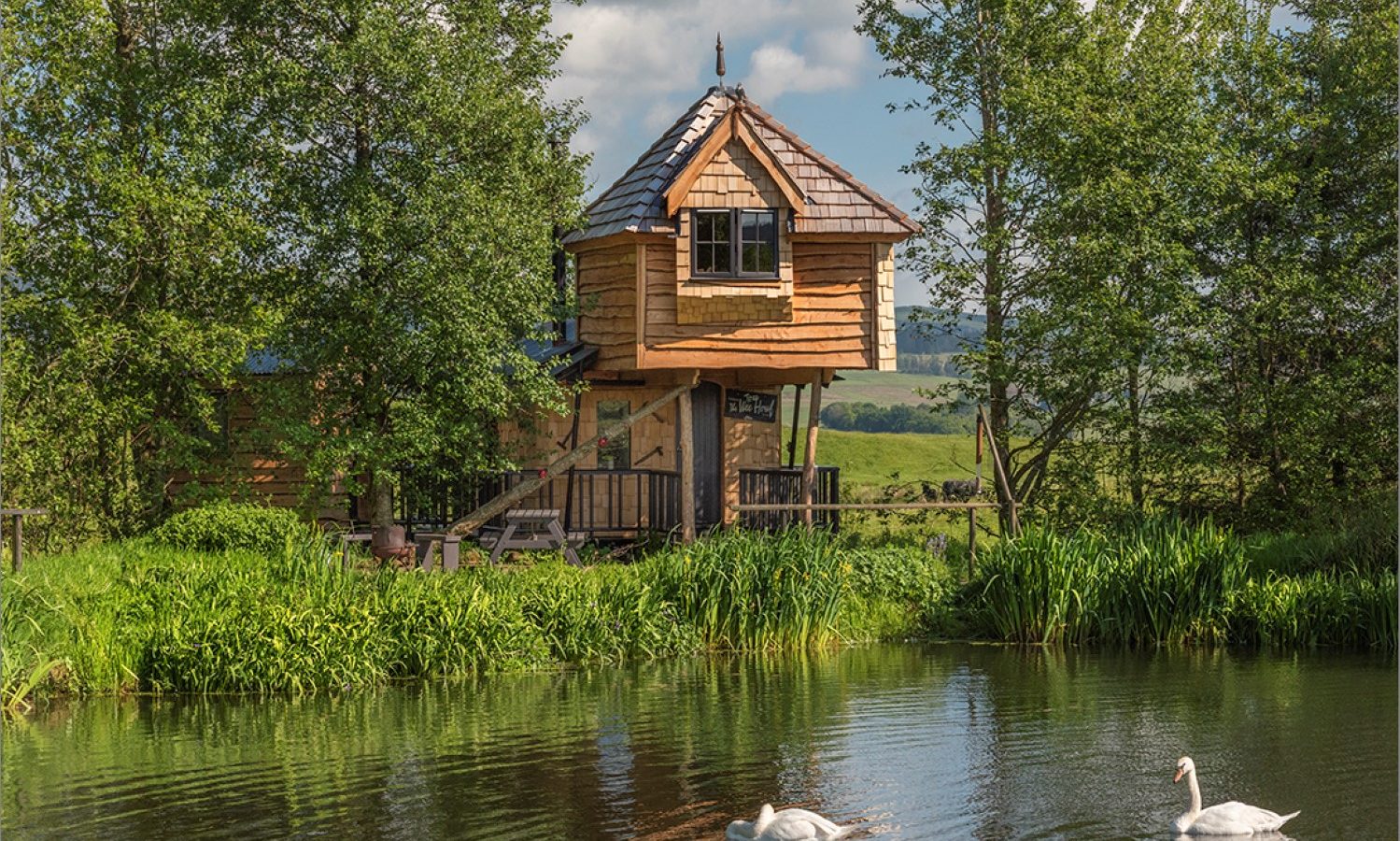 The charming wooden treehouse at Craighead Howf overlooks a serene pond with two graceful swans gliding by. It's enveloped in lush greenery and towering trees, all beneath a crystal-clear sky.