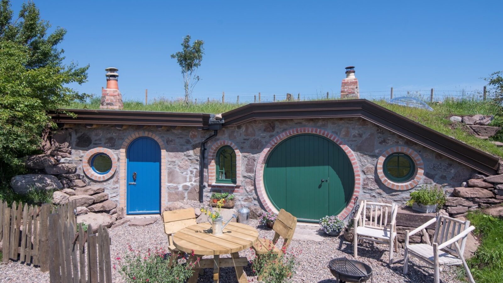 Discover the charm of a Craighead Howf-inspired hobbit-style house, featuring quaint round doors and chimney stacks. Relax in the small outdoor seating area, complete with a wooden table and chairs, perfect for enjoying tranquil moments surrounded by nature.