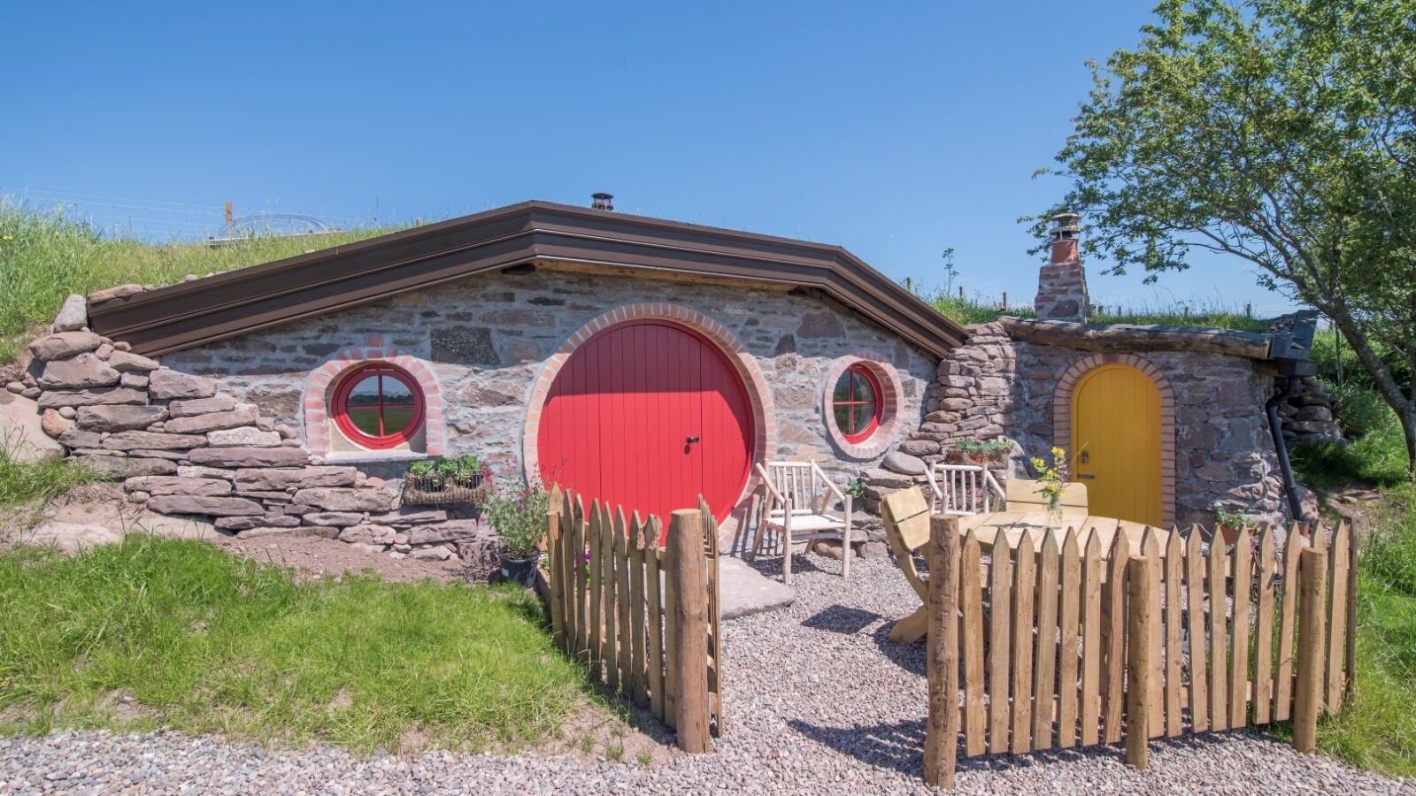 The stone and earth house at Craighead Howf features charming round red and yellow doors, encircled by a rustic wooden fence and lush trees, all beneath a clear blue sky.