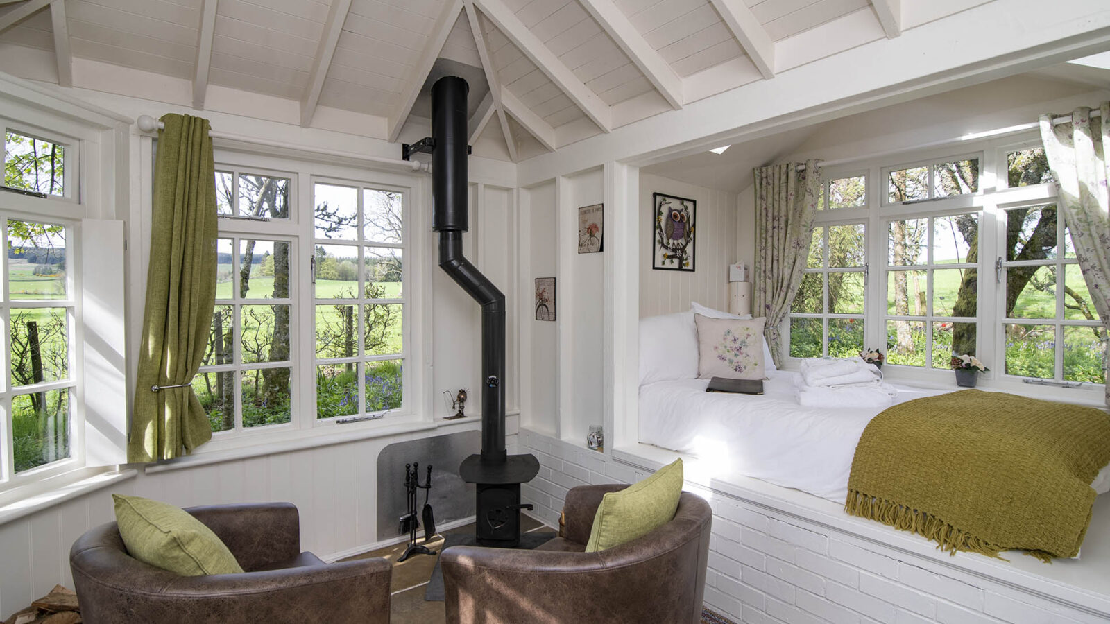 Cozy room at Craighead Howf with a wood-burning stove, two chairs, and a bed nestled by windows overlooking lush greenery. The space boasts white walls and ceiling.