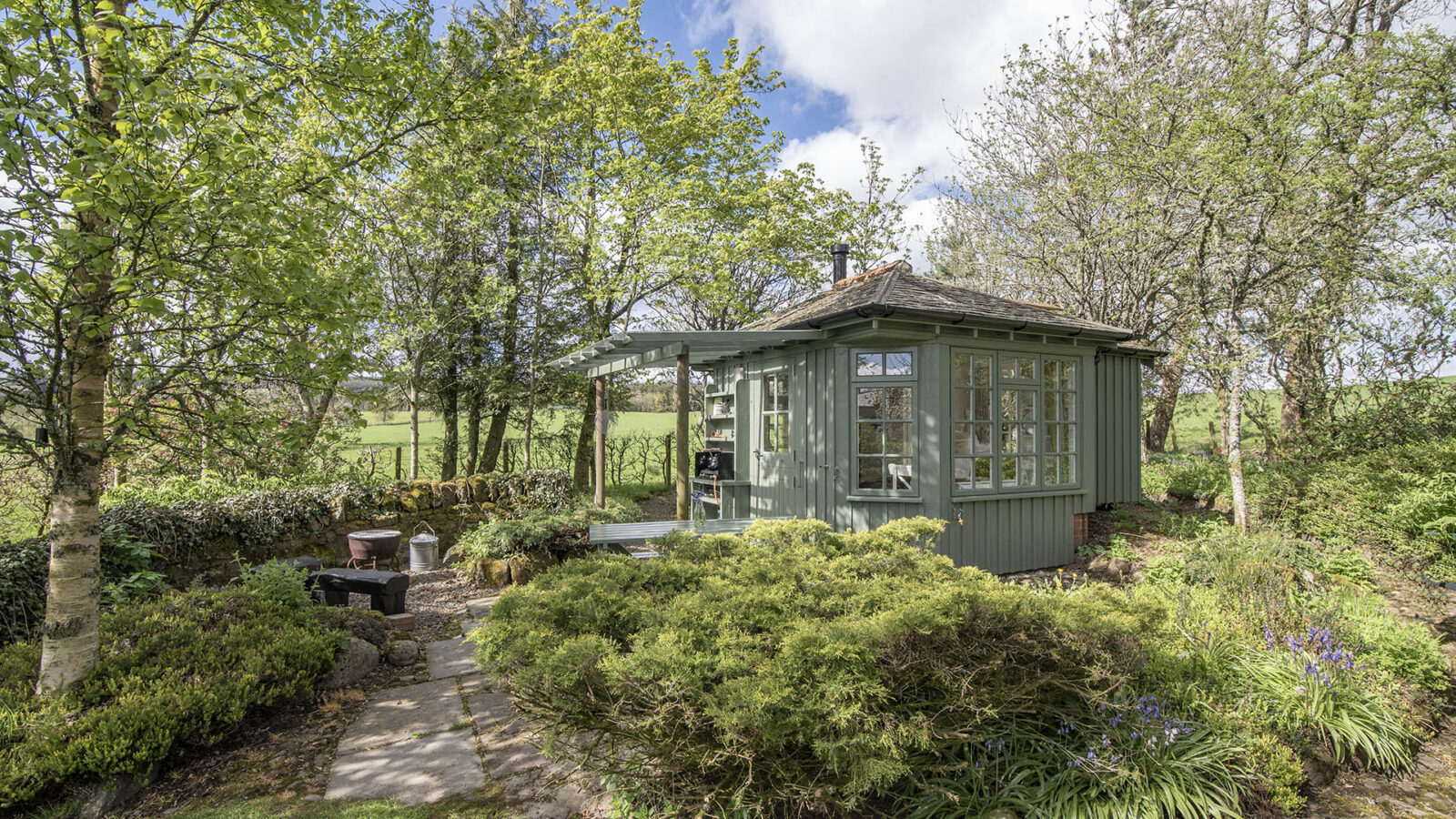 Nestled in the serene embrace of nature, this green wooden garden shed by Craighead Howf is surrounded by lush trees and shrubs. A charming stone pathway guides you to its door, promising a tranquil escape or unique accommodation on a sunlit day.