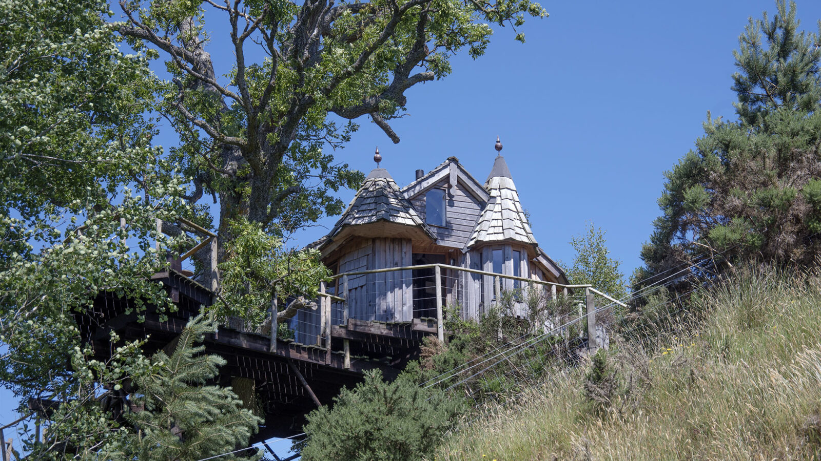 The Craighead Howf wooden treehouse, with its charming conical roofs, is nestled among trees on a serene hillside, set against a backdrop of clear blue sky.