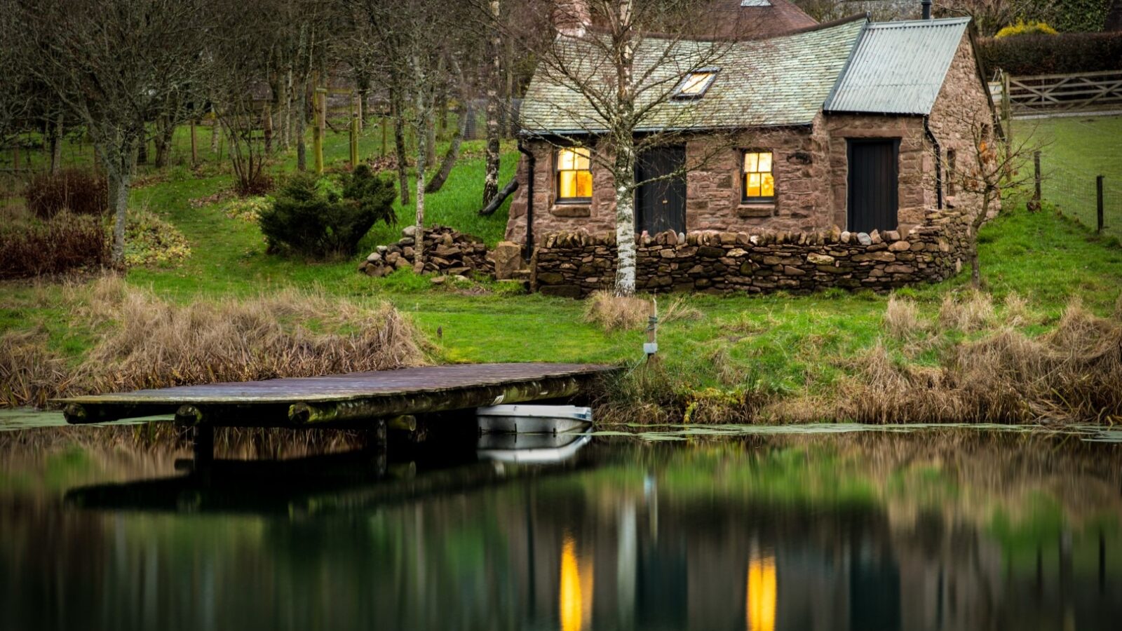 Nestled in the serenity of Craighead Howf, a cozy cottage with lit windows stands by a tranquil pond, surrounded by trees and lush greenery, perfectly reflecting the enchanting scene on the water's surface.