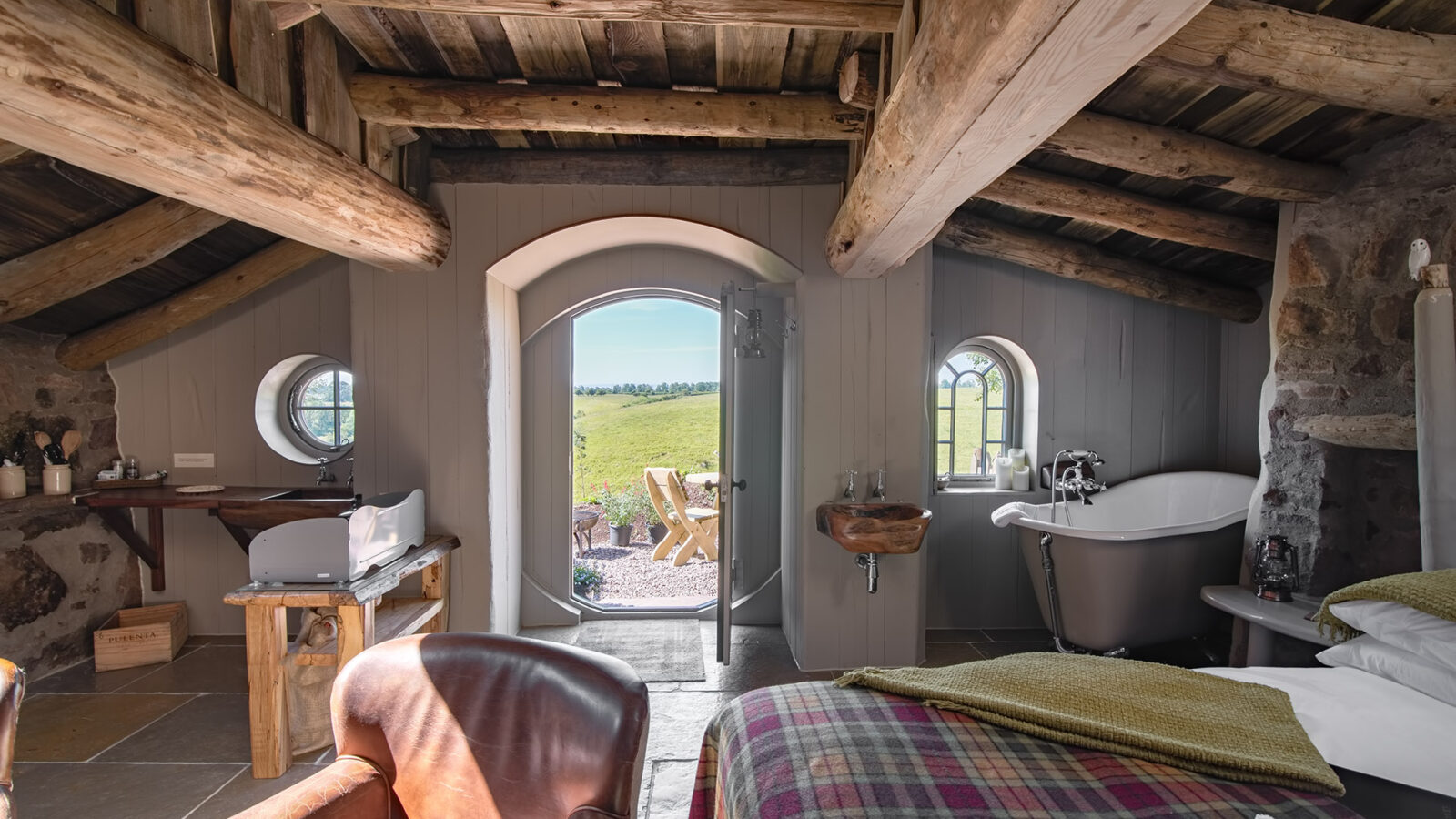 Cozy rustic room at Craighead Howf with wooden beams, a leather chair, bed, bathtub, and an open door leading to a grassy field outside.