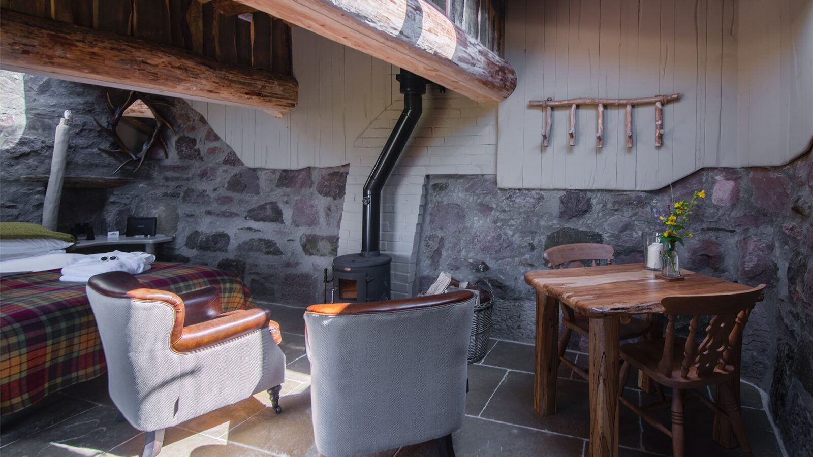 Experience the charm of a cozy rustic room at Craighead Howf, featuring stone walls, plush armchairs, and a wood-burning stove. Gather around the wooden table adorned with a vase of flowers for an unforgettable stay.