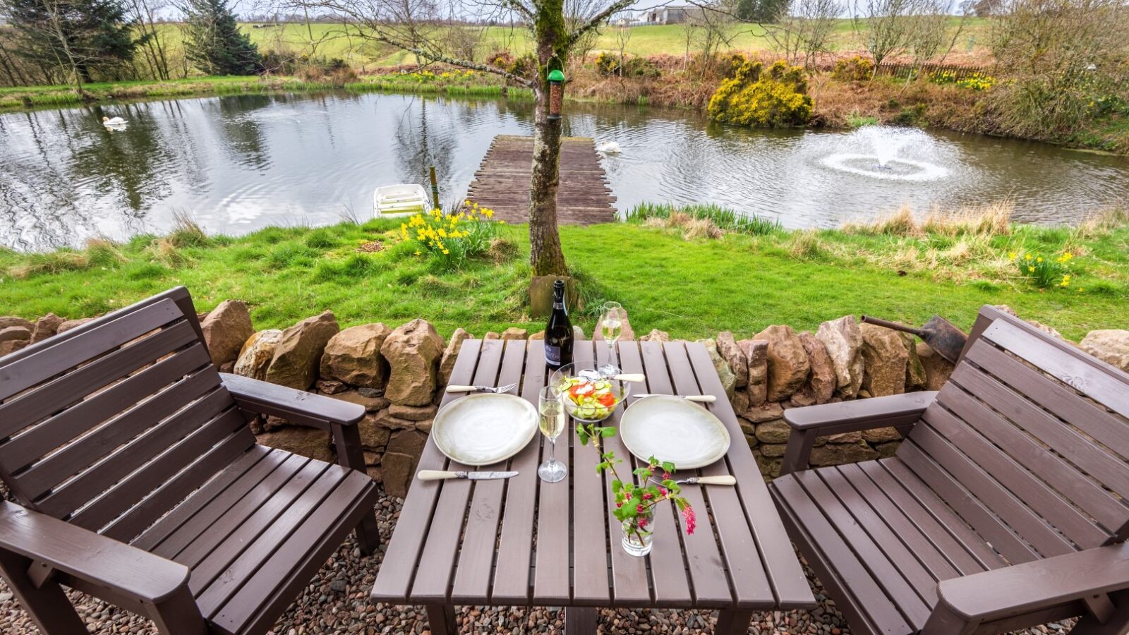 Two wooden chairs and a table with a meal and wine, overlooking a pond with a fountain and surrounded by greenery and trees—a perfect retreat in the heart of Craighead Howf, Scotland's charming countryside accommodation.