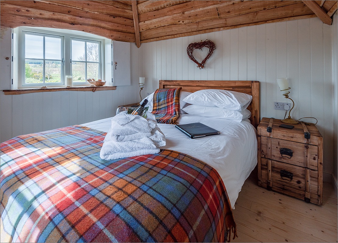 This cozy Craighead Howf bedroom features a wooden ceiling, a plaid blanket on the bed, heart-shaped wall decor, and a scenic window view.