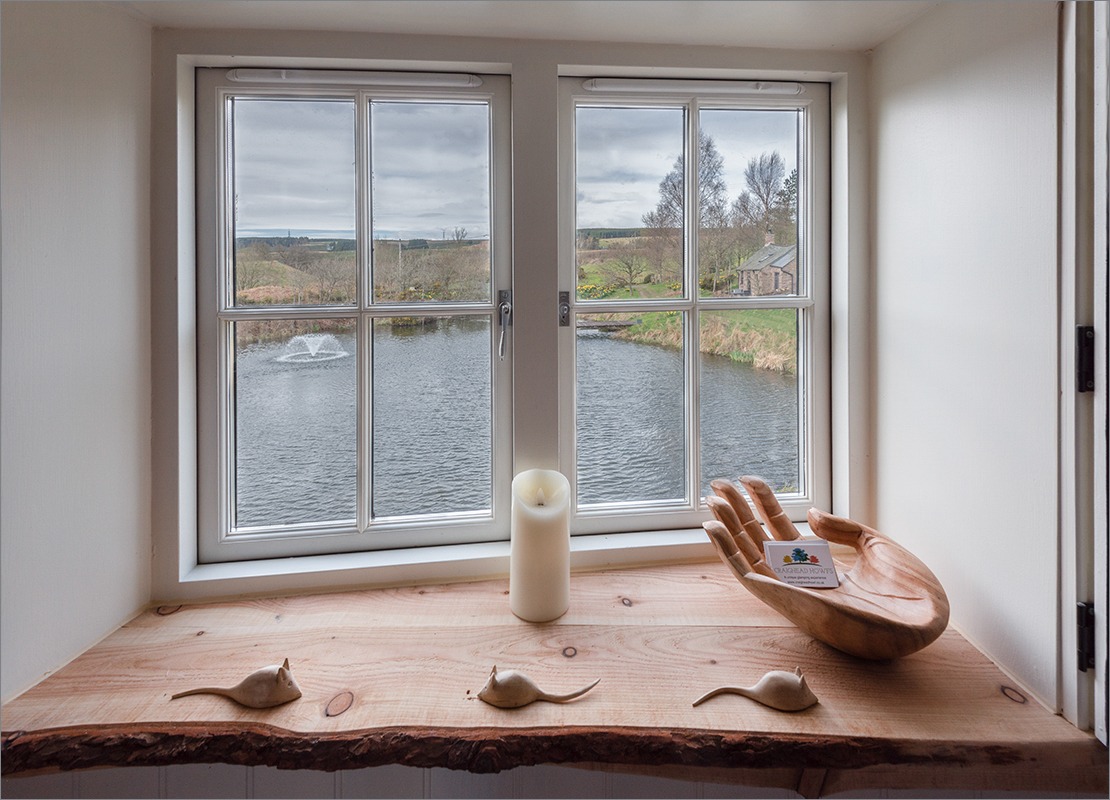 Through the window, a pond with a fountain is nestled among the Scottish countryside's rolling hills. On the windowsill, a candle, wooden hand, and stone mice add charm to this unique accommodation at Craighead Howf.