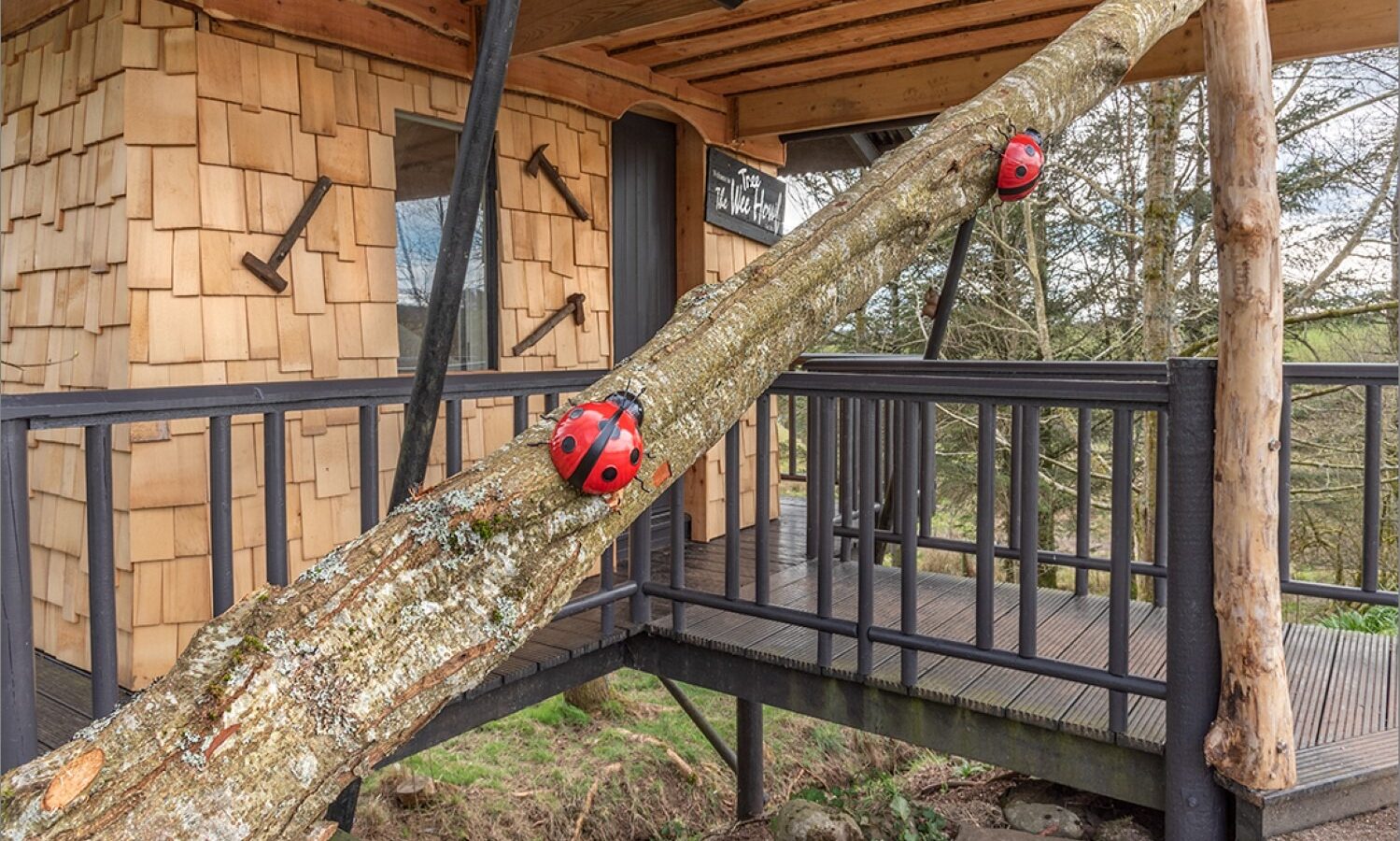 Nestled among the scenic landscapes of Scotland, this charming treehouse features wooden siding and two large red and black ladybug decorations adorning a slanted tree trunk. As part of the unique holiday accommodation at Craighead Howf, it promises an enchanting escape into nature.