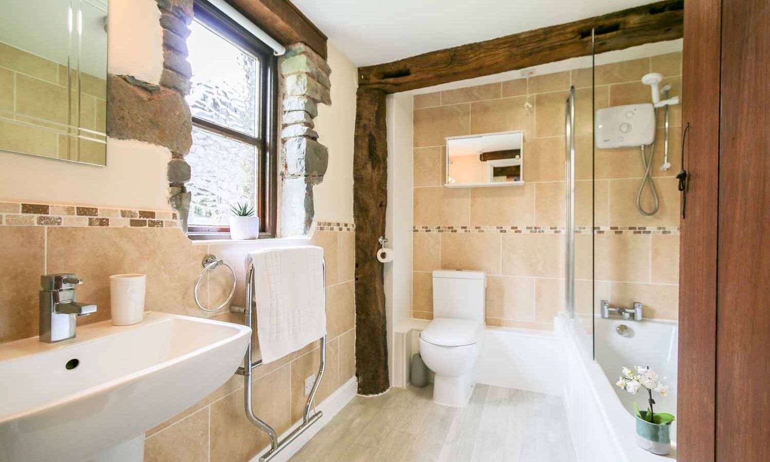 A modern bathroom at Cwm Chwefru Cottages with beige tiles and wood accents. It features a white sink with a silver faucet, a toilet, a bathtub with a glass shower screen, and a towel rack with a white towel. Natural light streams through the window, highlighting the decorative plants and flowers.