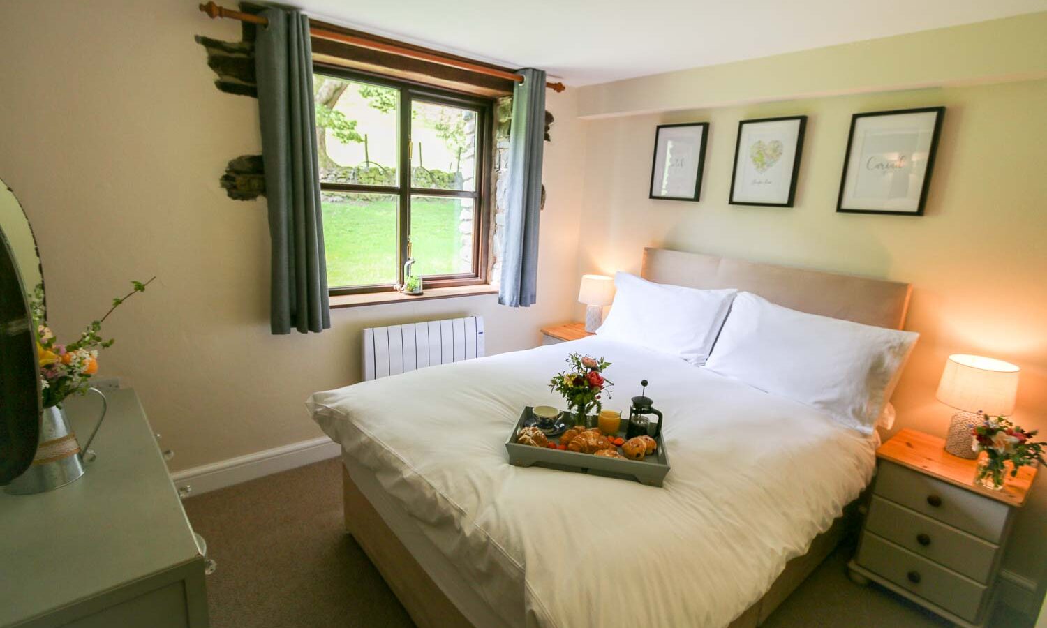 A cozy bedroom at Cwm Chwefru features a neatly made bed with white linens, complemented by a breakfast tray holding flowers, croissants, and a teapot. Two bedside tables with lamps flank the bed. The room's window has gray curtains, and framed artwork hangs above the bed, adding to its charming cottage feel.