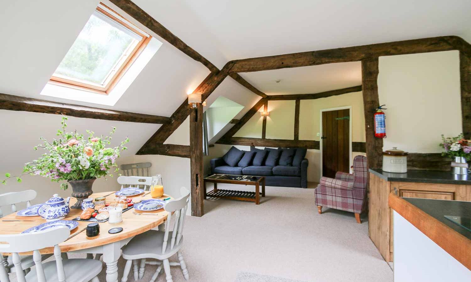 A cozy, rustic living area with exposed wooden beams, a skylight, and white walls. The room features a dining table set for breakfast, a plush gray sofa, and a checkered armchair. A bouquet of flowers sits on the dining table at Cwm Chwefru Cottages, enhancing the inviting atmosphere.