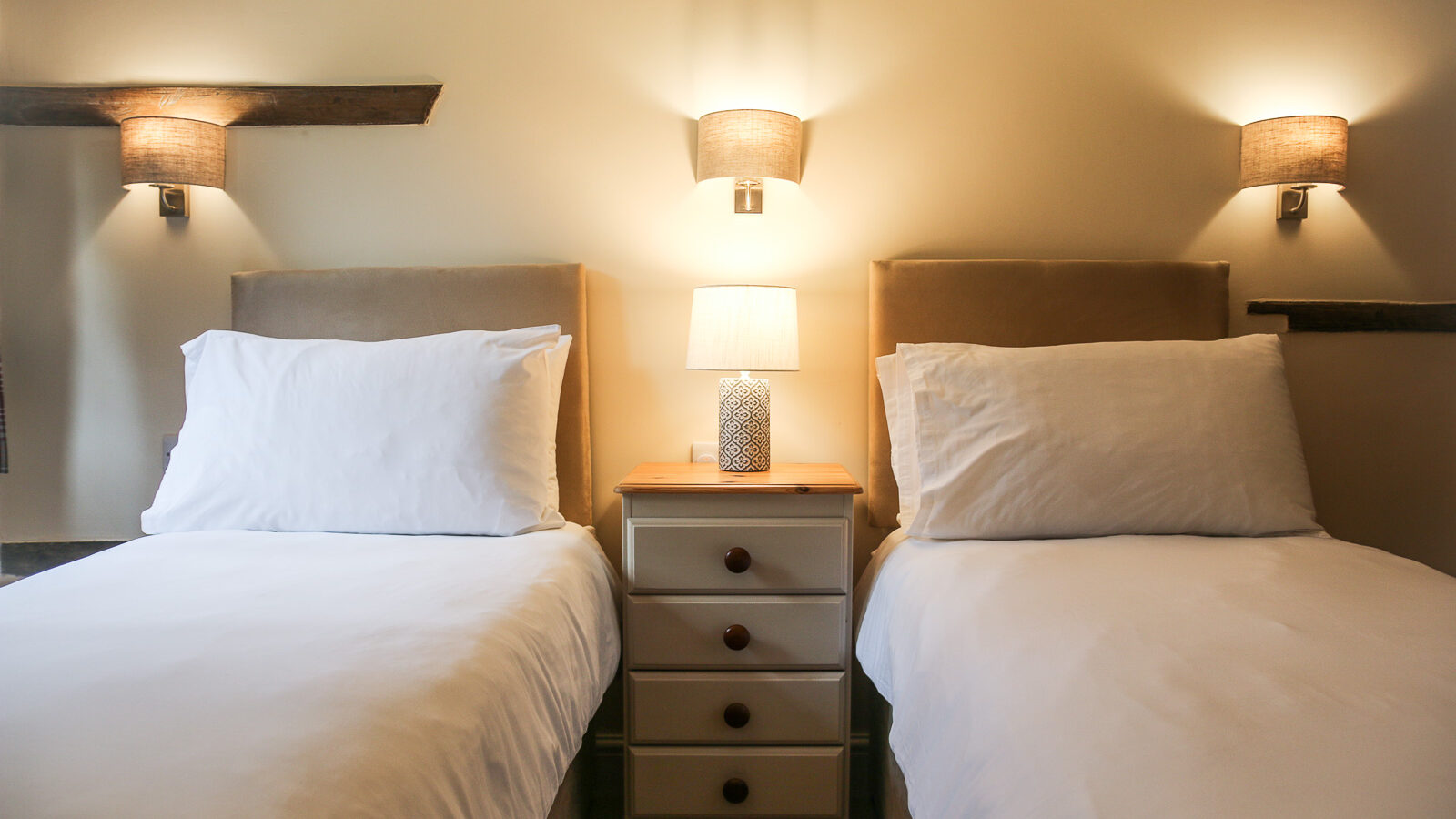 A cozy bedroom in Cwm Chwefru cottages features two single beds with crisp white bedding and pillows. A small wooden nightstand with a textured-base lamp sits between the beds, while two wall-mounted lamps with beige shades cast a gentle glow above.