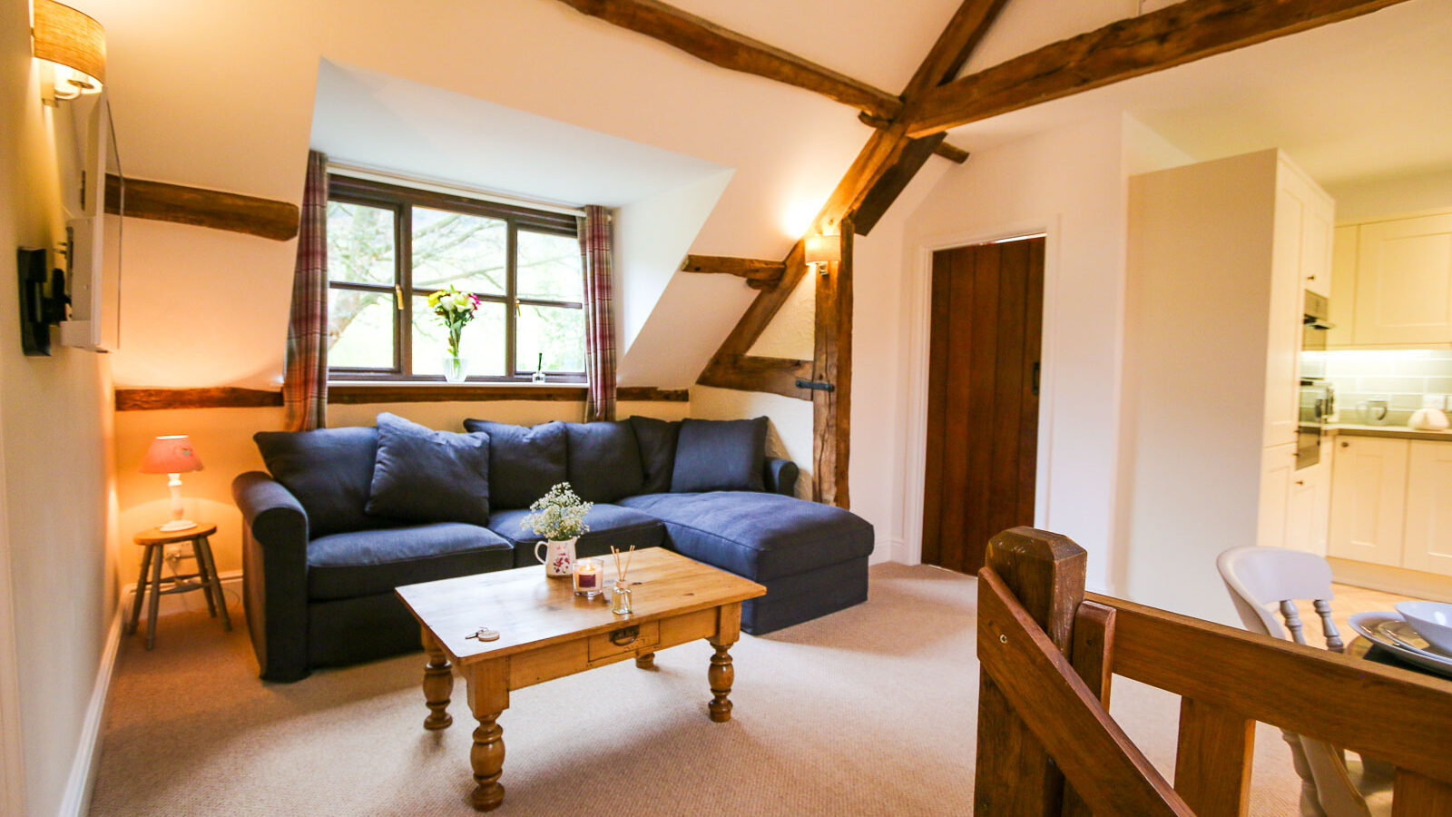 A cozy living room with wooden beams and slanted ceilings, centered around a blue sectional sofa. A wooden coffee table holds a small plant and decor items. A window with plaid curtains overlooks greenery, reminiscent of charming Cwm cottages. A door leads to an adjacent white kitchen area.
