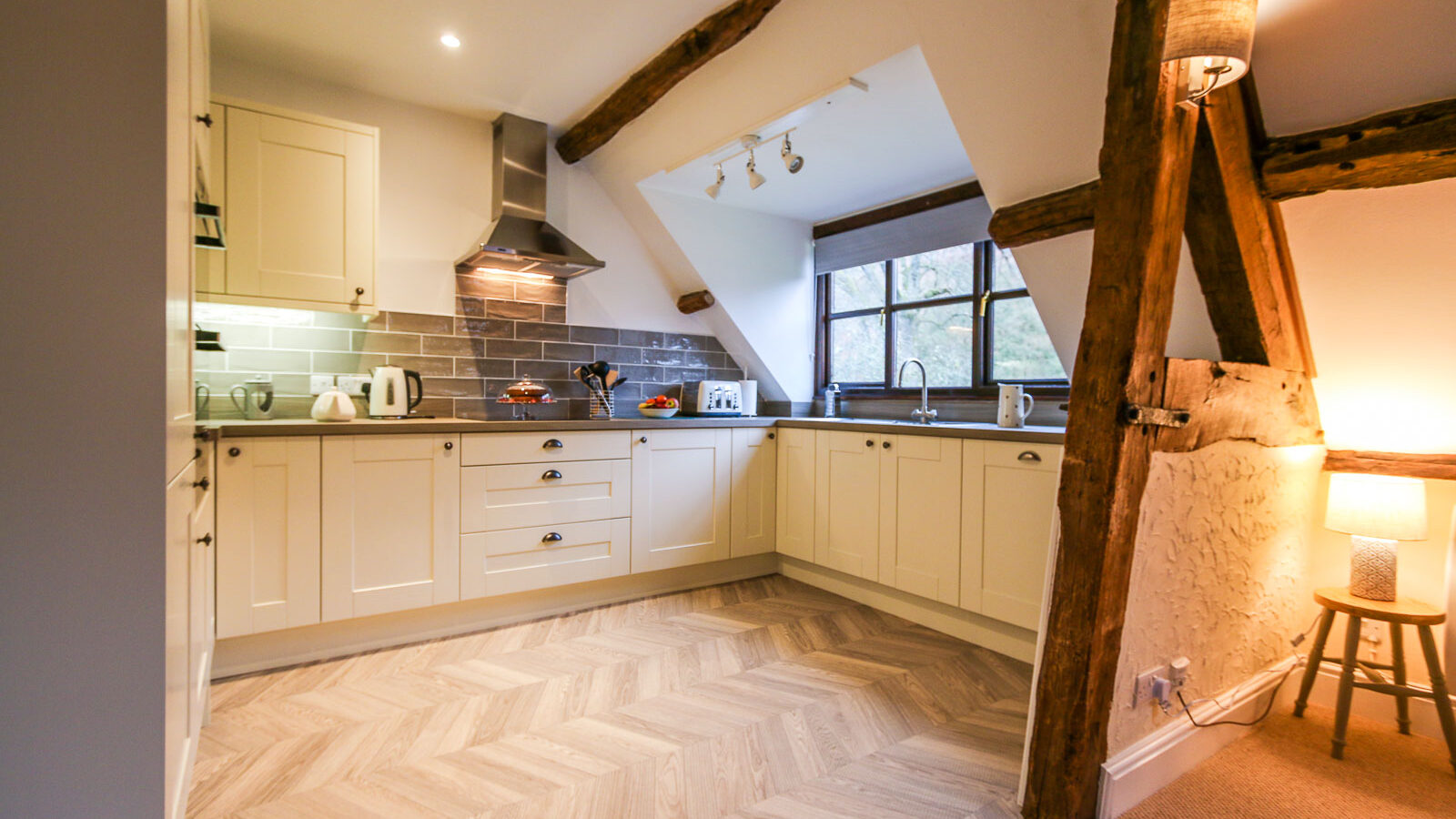 A cozy kitchen with rustic charm in one of the Cwm Chwefru Cottages, featuring white cabinetry, a tiled backsplash, a wooden beam ceiling, and a large window letting in natural light. The kitchen has modern appliances, including a kettle and toaster, with a wooden stool and lamp in the corner.