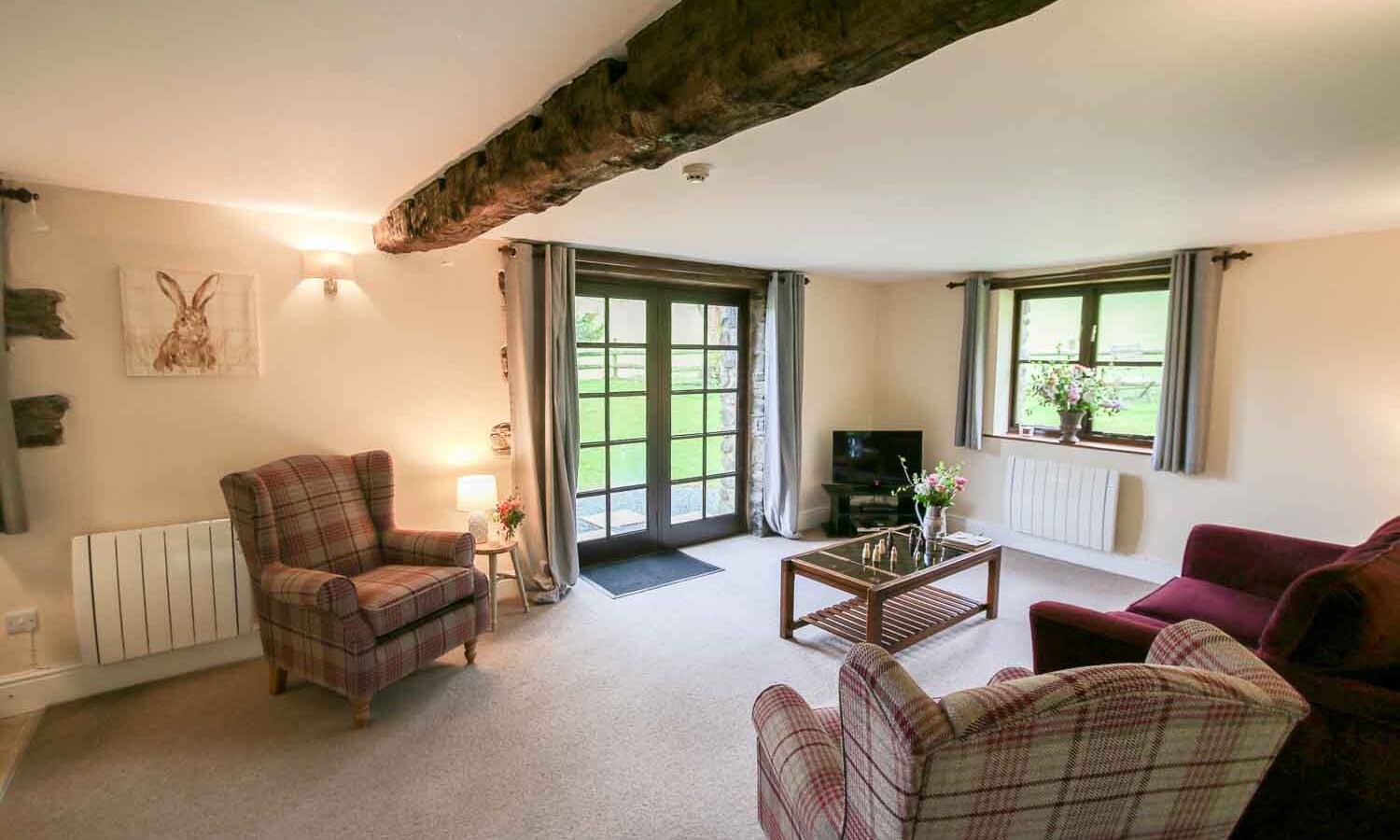 A cozy living room at Cwm Chwefru Cottages features two plaid armchairs, a maroon sofa, a coffee table with flowers, and a small TV. The room boasts a wooden beam on the ceiling, light walls, and large windows with curtains. French doors open to a lush outdoor space.