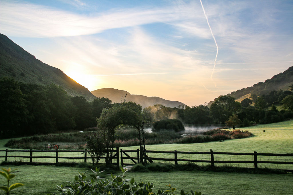 A serene landscape at sunrise with mist rising from a pond in the middle of a green field near Cwm Chwefru Cottages. The scene is framed by trees and hills, with a wooden fence in the foreground. The sky is streaked with light clouds and contrails from passing aircraft.