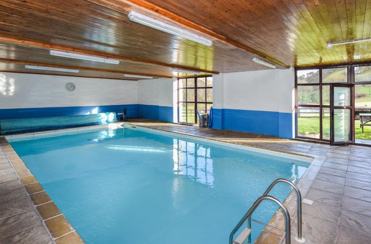 An indoor swimming pool with clear blue water, surrounded by a tiled floor. Large windows on one side let in natural light, complementing the wooden ceiling adorned with fluorescent lights. A ladder at the pool's edge offers easy access, and doors lead to the serene surroundings of Cwm Chwefru Cottages.