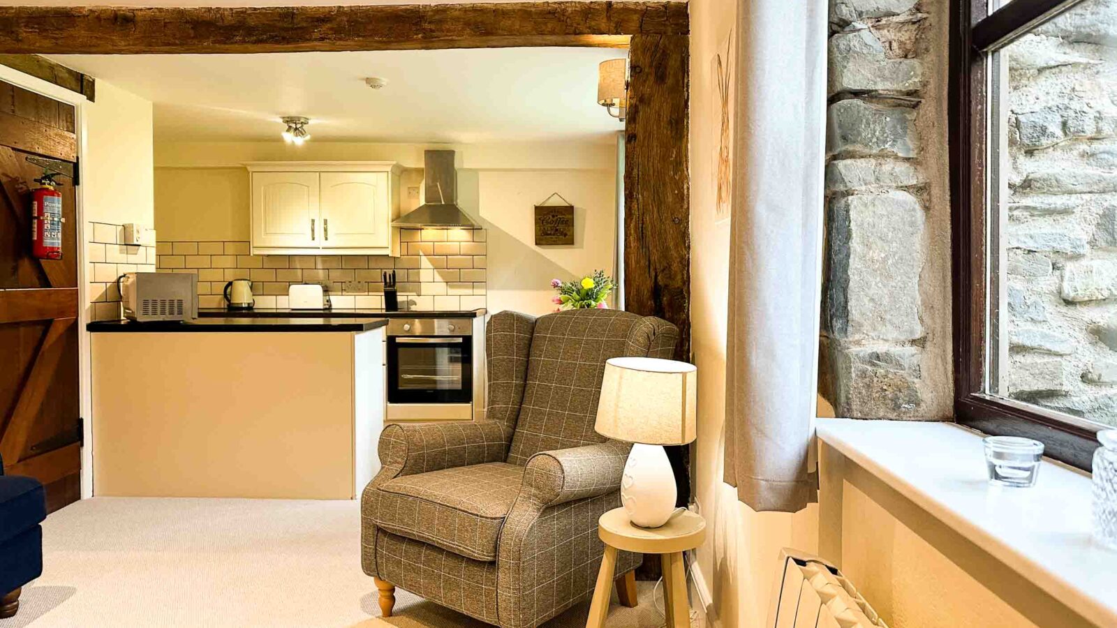 A cozy living area featuring a cushioned armchair beside a small wooden table with a lit lamp. The room has a rustic feel with exposed wooden beams and a stone wall next to a window, reminiscent of Cwm Chwefru cottages. The open kitchen has white cabinets and modern appliances, including a stainless steel oven.