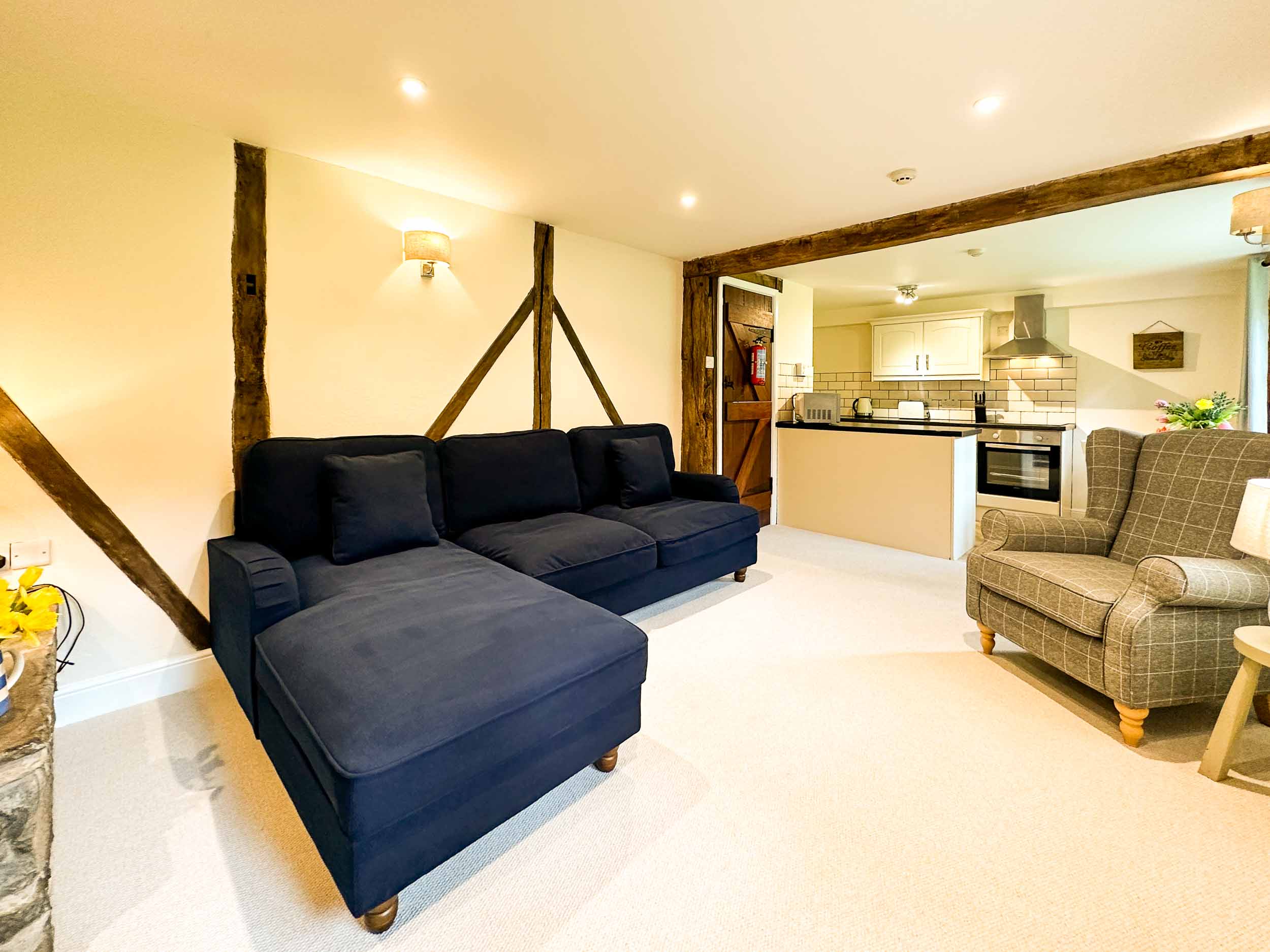 A cozy living room with exposed wooden beams features a large dark blue sectional sofa and a checkered armchair. In the background, there's an open-concept kitchen with light-colored cabinets, stainless steel appliances, and a small dining area, reminiscent of the charm found in Cwm Chwefru Cottages.