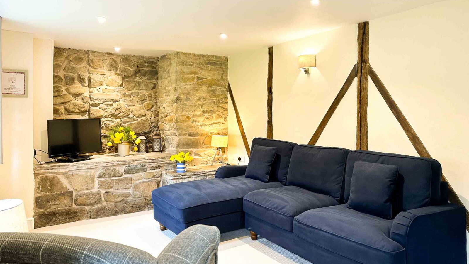 A cozy living room with a stone accent wall, a navy blue sectional sofa, a checkered armchair, and a TV. Reminiscent of charming cottages like those in Cwm Chwefru, the room is well-lit with recessed lighting and a wall sconce. Decorative items and flowers adorn a shelf below the TV.