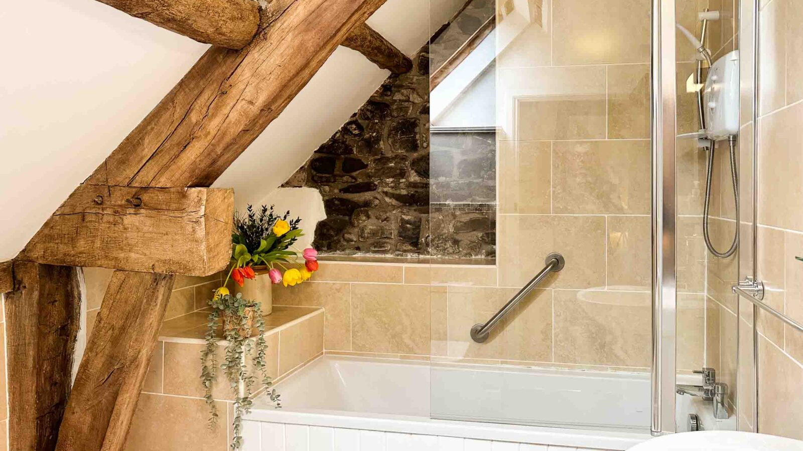 A cozy attic bathroom in Cwm Chwefru cottages featuring exposed wooden beams and rustic stone walls. The space includes a modern bathtub with a glass shower screen and a mounted showerhead. A vase with colorful flowers adds a touch of greenery and vibrancy to the serene setting.