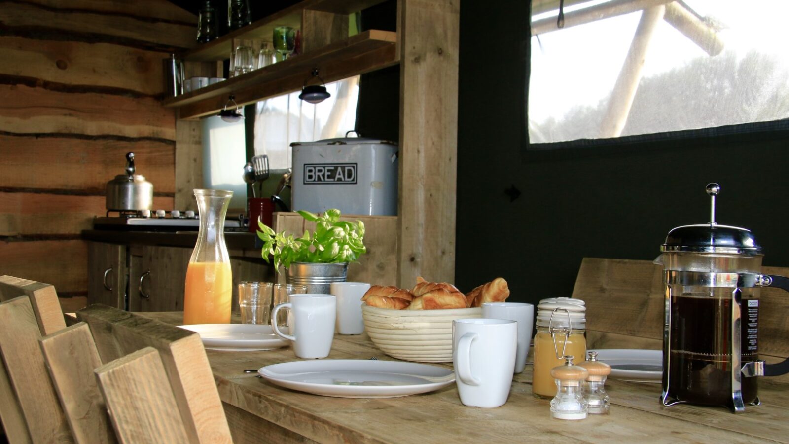 Rustic kitchen at Sloeberry Farm, with a wooden table set for breakfast. It features croissants, juice, coffee, and a bread box on shelves—perfectly capturing the charm of rural lodges.