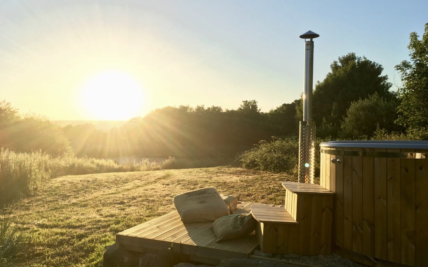 A wooden hot tub with steps graces the grassy landscape at Sloeberry Farm, under a clear sky with the bright sun on the horizon—a perfect retreat near charming lodges.