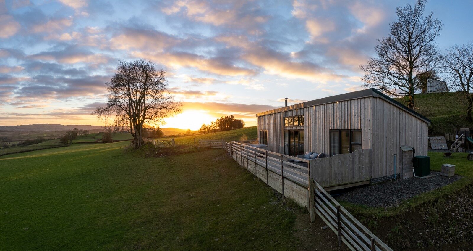 Nestled on a hillside, Penlan Lodges offer a charming wooden cabin with a sprawling grassy lawn, all bathed in the warm hues of a sunset sky dotted with scattered clouds. This idyllic accommodation provides the perfect retreat for those seeking tranquility and natural beauty.