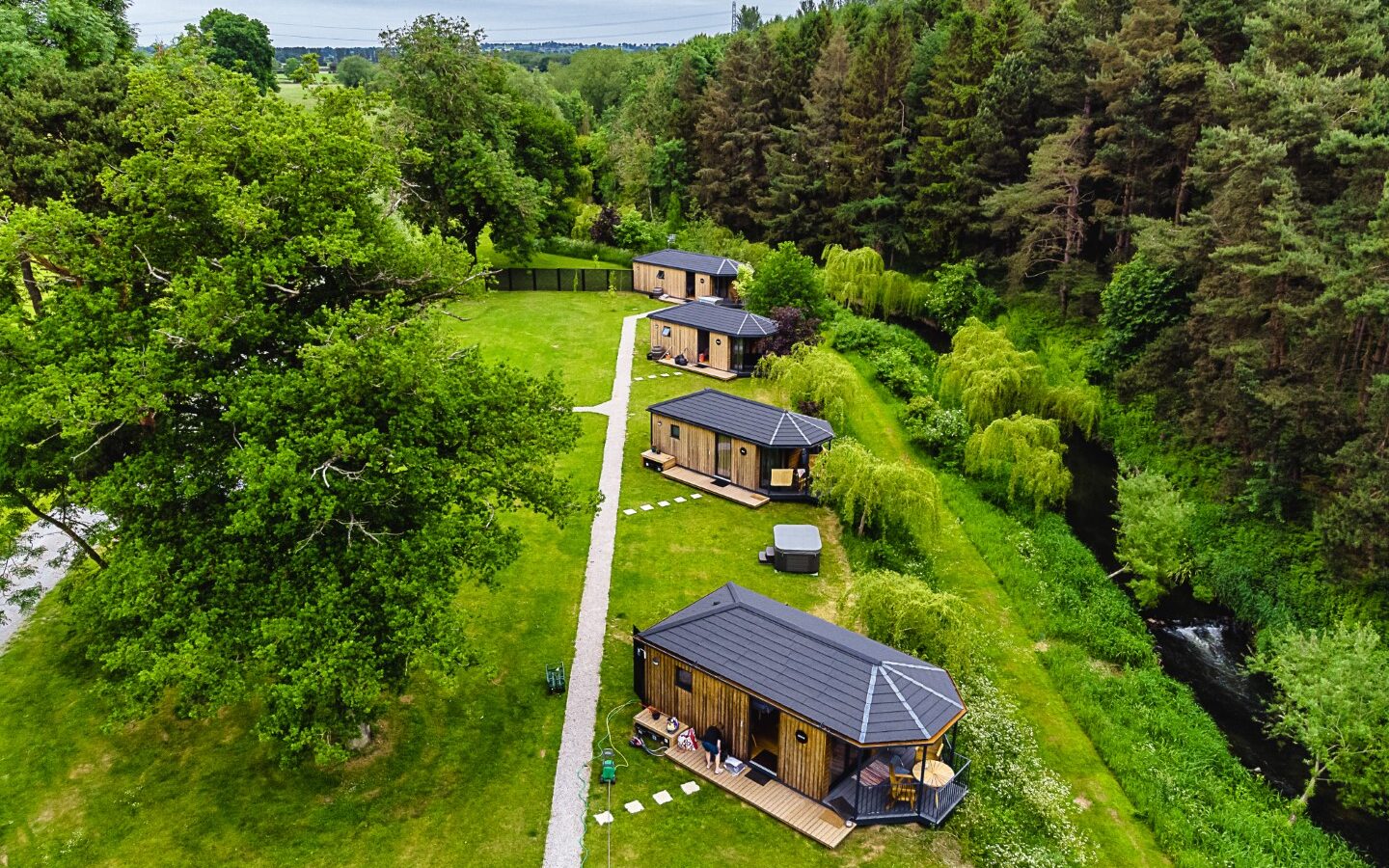 Aerial view of four cabins lining a riverside path, surrounded by lush green grass and towering trees.