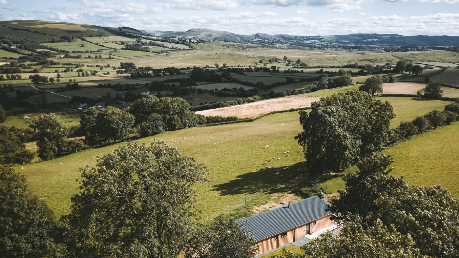 An aerial view of a rural landscape with rolling hills, fields, and trees surrounding Penlan Lodges under a cloudy sky.