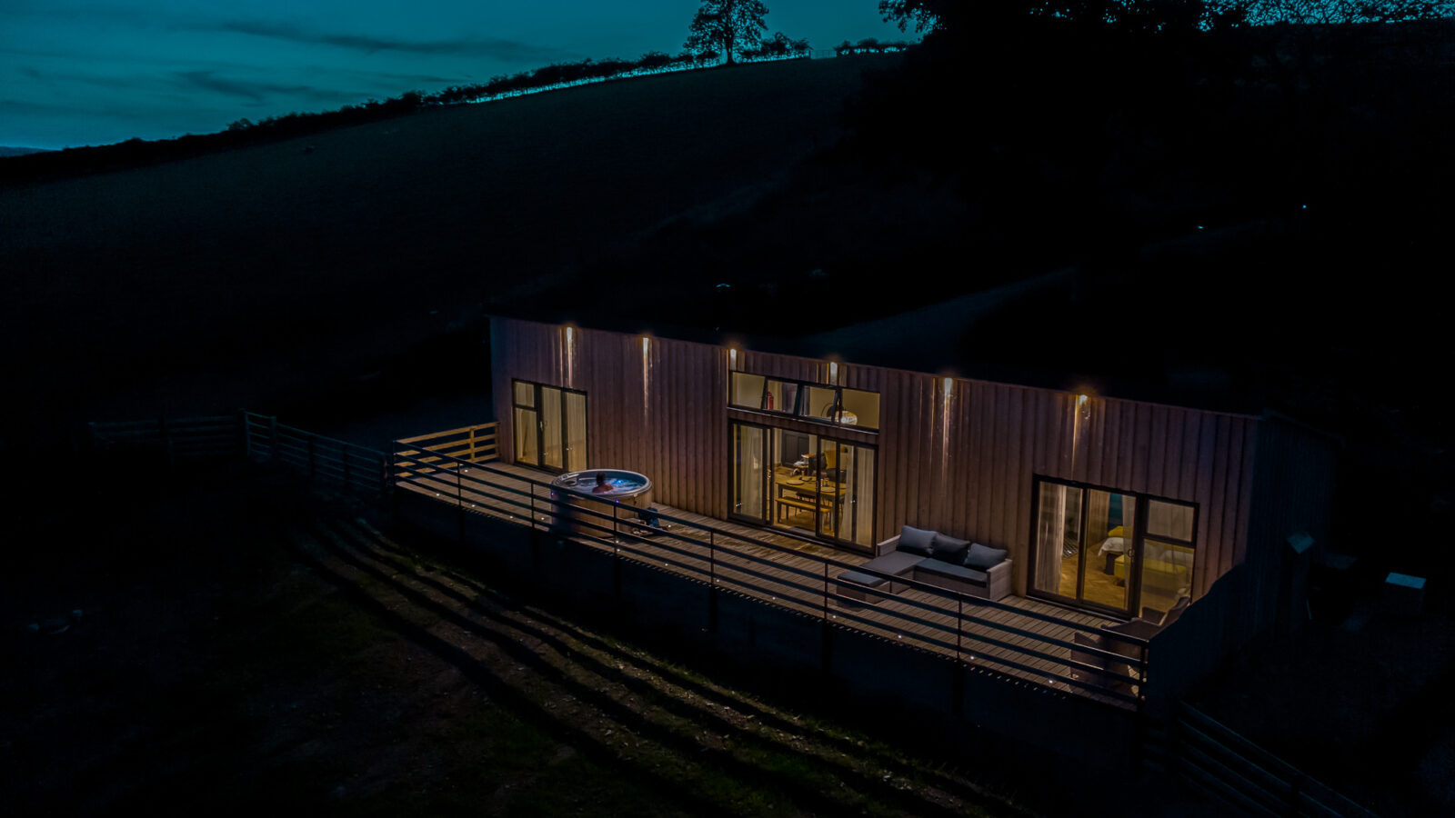 The modern Penlan Lodges cabin boasts large windows and outdoor seating, glowing softly against the hillside at night.