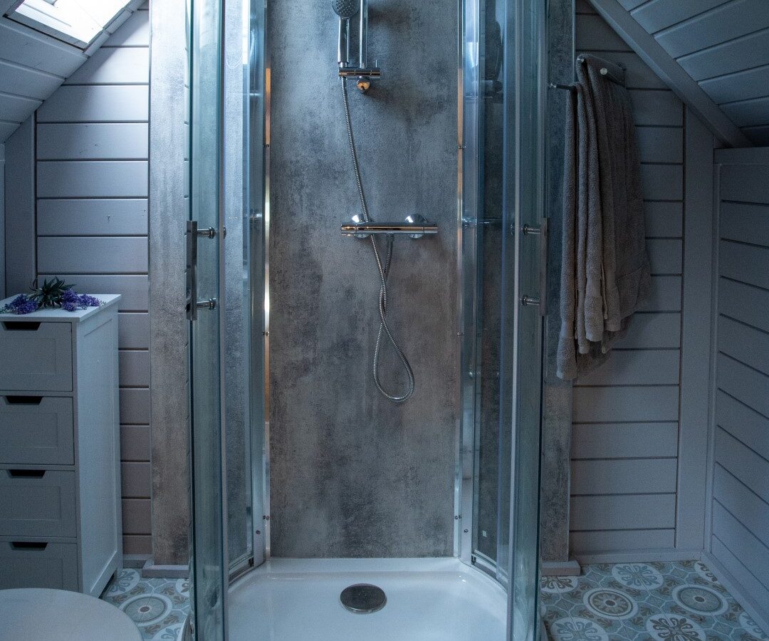 A sleek glass shower enclosure graces the attic bathroom, its modern elegance accentuated by gray walls and patterned floor tiles. A skylight casts light across the room, creating an ethereal atmosphere reminiscent of a Dragon's Gate hidden in a serene hideaway.