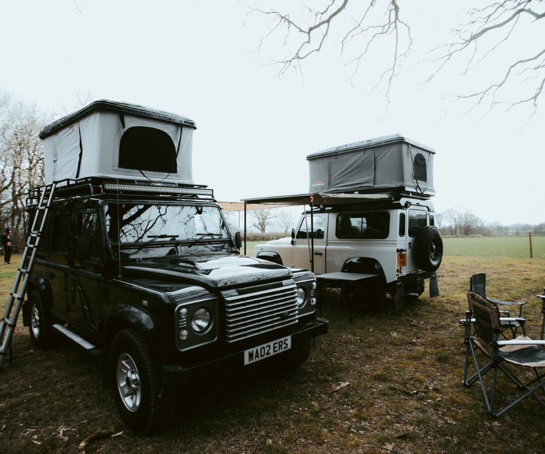 Two off-road vehicles, including a rugged Defender, with rooftop tents parked next to each other on grass, create a perfect camping scene with bare trees and camping chairs in the background.