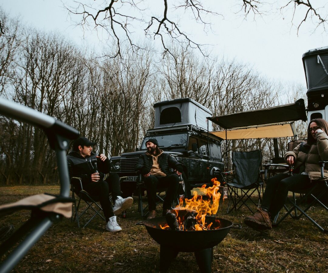 Three people sit around a campfire, enjoying a Defender camping experience near their parked SUV with a roof tent, nestled in a wooded area among bare trees.